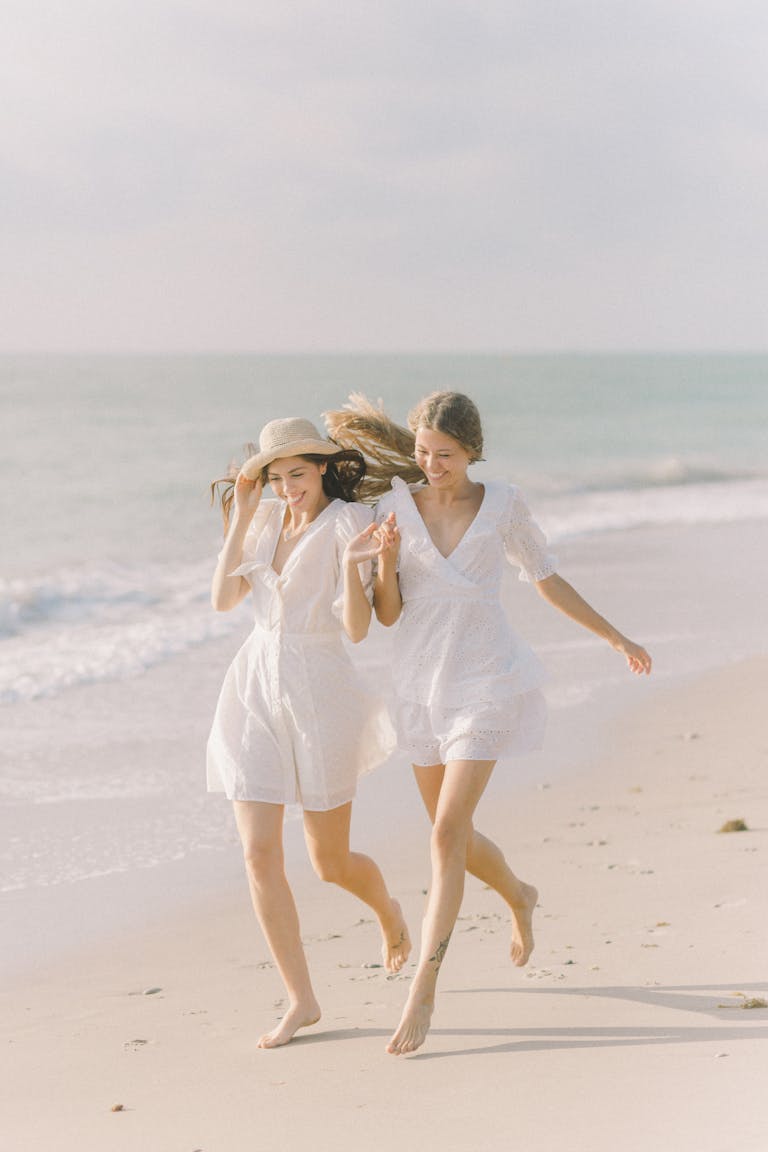 Two women in white dresses running happily along a sunny beach, enjoying summer vibes.